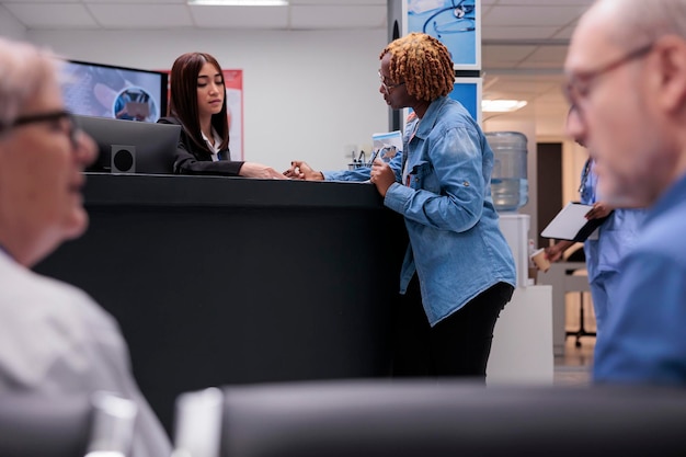 African american person filling in report files, talking to receptionist at hospital reception counter. Woman writing medical form before checkup appointment with doctor at health center.