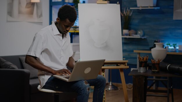 집에 있는 예술품 방에 앉아 새로운 영감을 얻기 위해 노트북을 사용하는 아프리카계 미국인. 현대 걸작 창작 및 그림을 위한 기술과 장치를 갖춘 흑인 젊은 예술가