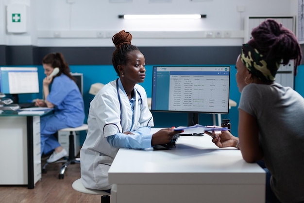 Афро-американский врач обсуждает анкету с симптомами болезни с больным пациентом во время клинического приема, работающего в офисе больницы. Практикующий врач, объясняя медицинское лечение