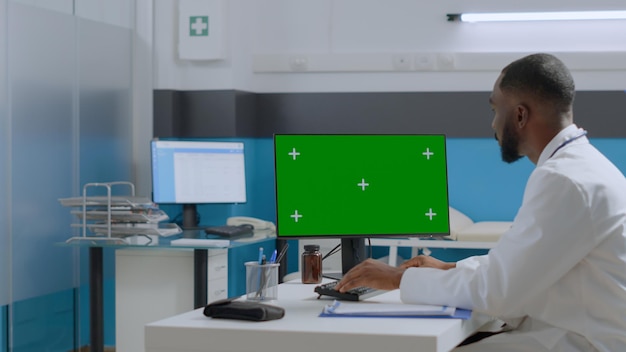 医療の専門知識を入力する分離されたディスプレイを備えたモックアップグリーンスクリーンクロマキーコンピューターを見ているアフリカ系アメリカ人の医師。病院のオフィスで病気のレポートを分析するセラピストの男性