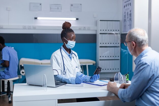 African american physician expert holding vial of covid medicine while talking to senior man about health risks. Clinic specialist doctor conversating with retired man about virus pandemic outbreak