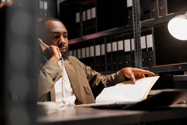 Free photo african american police investigator answering landline phone call in office at night time. concerned detective discussing crime case insight while analyzing forensic report file