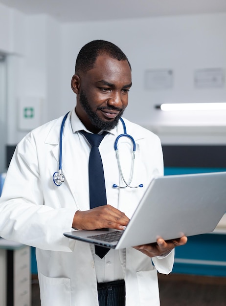 African american practitioner doctor holding laptop checking sickness symptoms analyzing medicine prescription for sick patient during medical appointment. Man working in hospital office