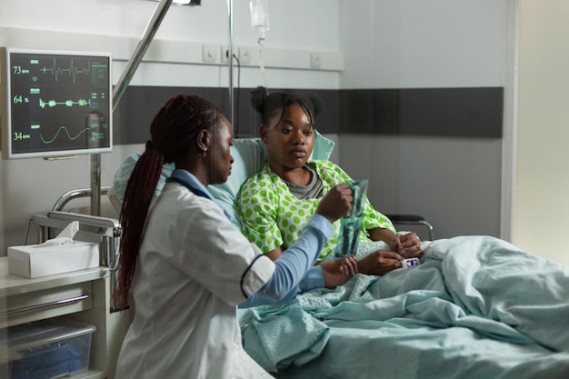 African american practitioner doctor standing beside sick young patient during medical appointment explaining bones xray expertise in hospital ward. Physician woman discussing healthcare expertise