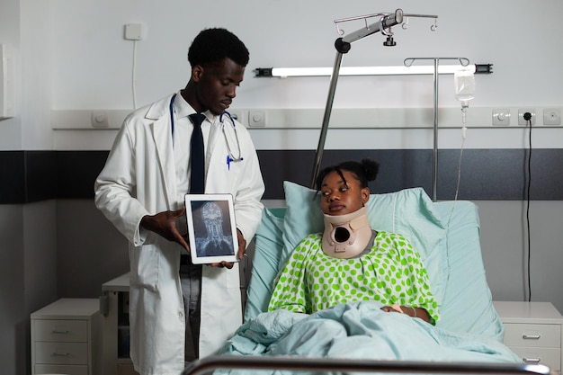 Афро-американский радиолог мужчина держит планшет, обсуждая рентгенографию костей с больной женщиной во время медицинского обследования в больничной палате. Пациентка в шейном воротнике восстанавливается после операции