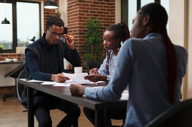 African american research agency coworkers in meeting about startup project financial status. Business people at desk in company office finance department talking about marketing expenses.