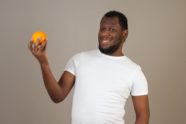 Афро-американский улыбающийся человек смотрит на мандарин в руке, стоя перед серой стеной.