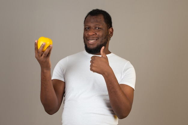 Uomo sorridente afroamericano con una mela cotogna in una mano e facendo il segno perfetto con l'altra mano, in piedi di fronte al muro grigio.