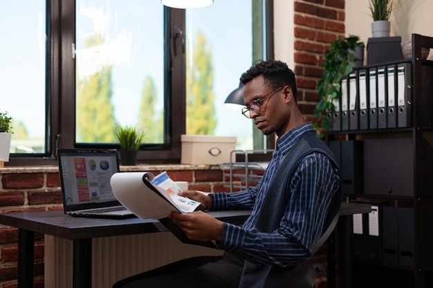 African american startup owner with glasses holding clipboard and paper with bussiness charts comparing data with sales results on laptop. Entrepreneur analyzing marketing indicators on laptop.