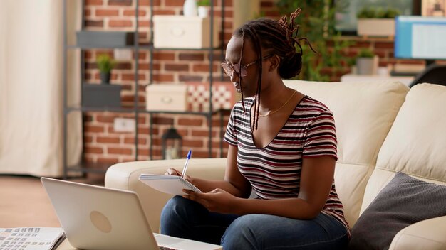 アフリカ系アメリカ人の学生がオンライン クラスのレッスンでメモを取り、ノートを使用してビジネス情報を書きます。ラップトップでインターネットを閲覧し、学校のウェビナーに参加する女性のフリーランサー。