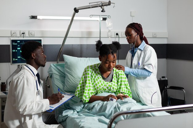 Афро-американский врач-терапевт, слушая легкие пациента с помощью стетоскопа, обсуждает медицинский опыт с терапевтом. Больная молодая женщина отдыхает в постели, выздоравливая после операции в больничной палате