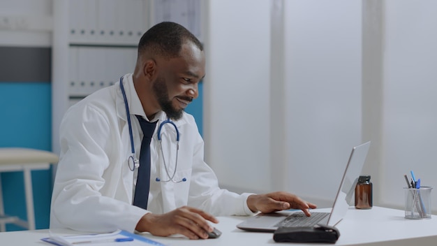 机に座っているアフリカ系アメリカ人のセラピスト医師は、病院のオフィスで医療治療で働いているラップトップの病気の専門知識を入力します。病気のレポートを分析する医師の男。医学の概念
