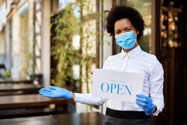 African American waitress holding open sign while wearing protective face mask and gloves at sidewalk cafe