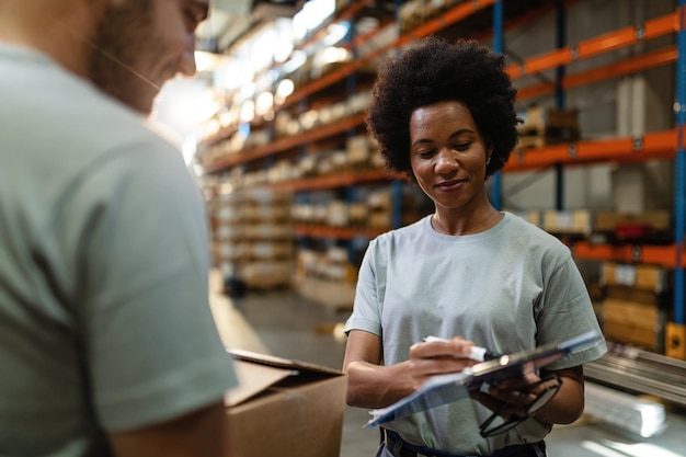 アフリカ系アメリカ人の倉庫作業員が、配送倉庫で作業しているときに、同僚に出荷用の製品のチェックリストを表示します