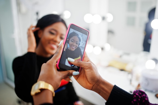 ビューティーサロンでメイクアップアーティストによるメイクをするアフリカ系アメリカ人女性アーティストが自分の作品の携帯電話で写真を撮る