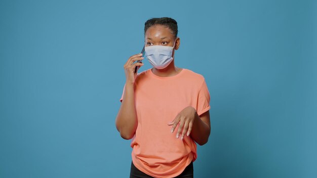 African american woman having conversation on phone call while wearing face mask against coronavirus. Young person using smartphone to talk to friends in studio during covid 19 pandemic.