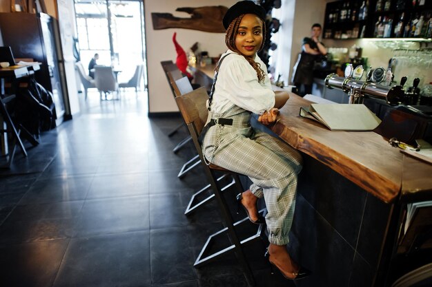 African american woman in overalls and beret sitting at bar counter