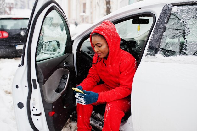 Африканская американка в красной толстовке с капюшоном сидит в машине в зимний снежный день с мобильным телефоном