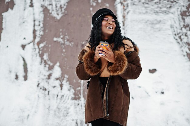 羊皮のコートとキャップのアフリカ系アメリカ人の女性が一杯のコーヒーと雪の背景に冬の日にポーズをとった