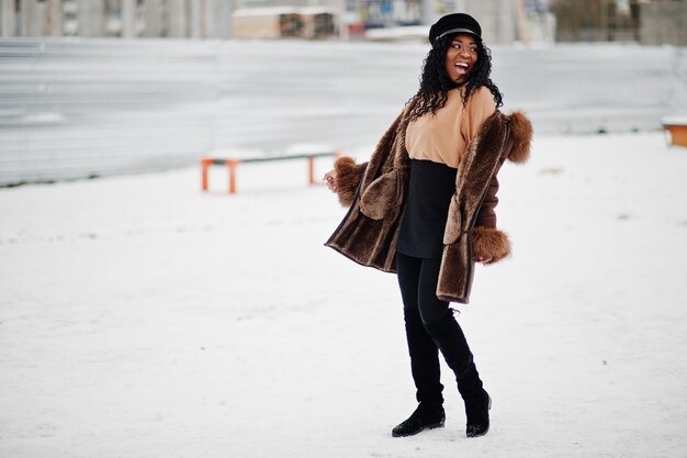 겨울날 눈 덮인 배경에 대해 포즈를 취한 양가죽 코트와 모자를 쓴 아프리카계 미국인 여성
