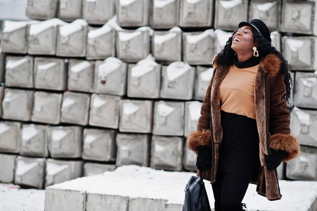 African american woman in sheepskin coat and cap posed at winter day against snowy stone background