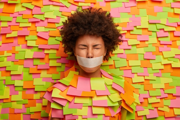 African American woman shut with taped mouth, forced to be silent, closes eyes, afraids of speaking, sticks out head in paper wall, colored stickers for writing information, has no freedom of speech