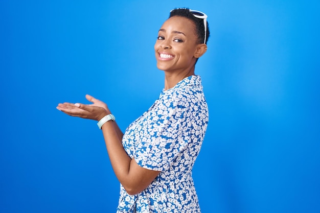 African american woman standing over blue background pointing aside with hands open palms showing copy space presenting advertisement smiling excited happy