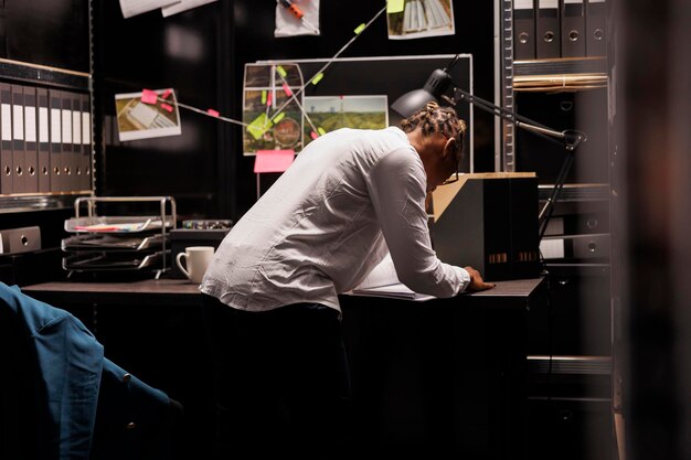 Free photo african american woman standing near detective desk and reading crime case file. police investigator studying forensic expertise material and searching investigation insight at night