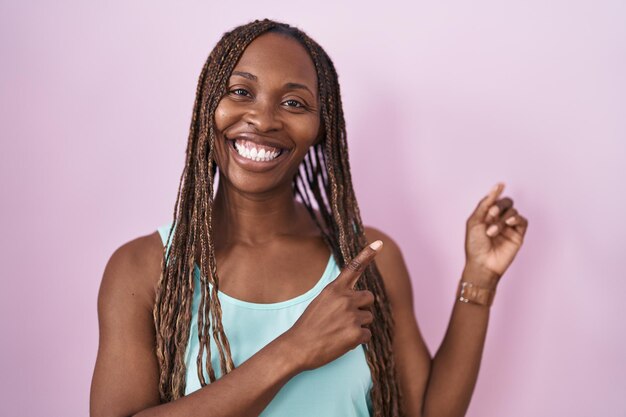 분홍색 배경 위에 서 있는 아프리카계 미국인 여성은 미소를 지으며 두 손과 손가락으로 옆쪽을 가리키는 카메라를 바라보고 있습니다.