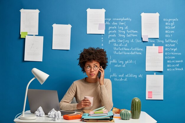 african american woman talks on mobile phone at workplace, chats with friend during coffee break, solves distance issues