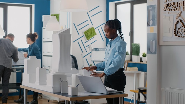 African american woman using laptop to design building model and blueprints plan. Engineer working on computer to create architectural project with urban construction structure.