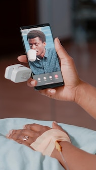 Donna afroamericana che utilizza smartphone con videochiamata per parlare con l'uomo mentre è seduto nel reparto ospedaliero. giovane paziente con sacca antigoccia iv che parla con un adulto in videoconferenza online.