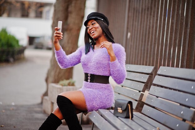 African american woman at violet dress and cap posed outdoor sitting on bench and making selfie on phone