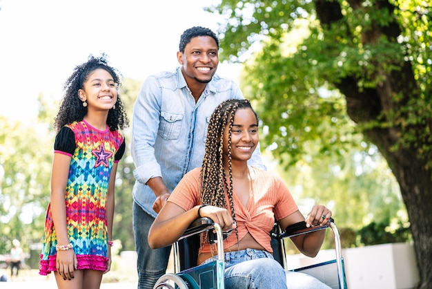휠체어를 탄 아프리카계 미국인 여성이 딸과 남편과 함께 야외에서 산책을 즐기고 있습니다.