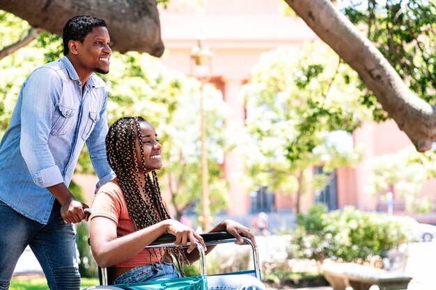 Афро-американская женщина в инвалидной коляске наслаждается прогулкой в парке со своим парнем.