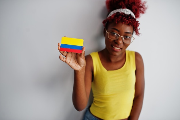 アフロの髪を持つアフリカ系アメリカ人の女性は黄色の一重項を着用し、眼鏡は白い背景で隔離のコロンビアの旗を保持します