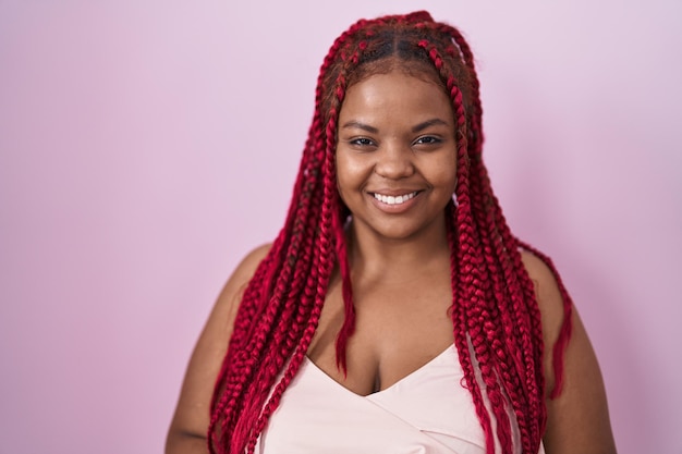 ピンクの背景の上に立って編んだ髪を持つアフリカ系アメリカ人の女性が手を合わせ、指を組んでリラックスした陽気な笑顔を浮かべています。成功と楽観的