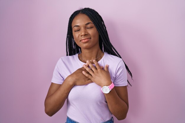 분홍색 배경 위에 머리를 땋고 눈을 감은 채 손을 가슴에 얹고 얼굴에 감사하는 몸짓으로 웃고 있는 아프리카계 미국인 여성. 건강 개념입니다.