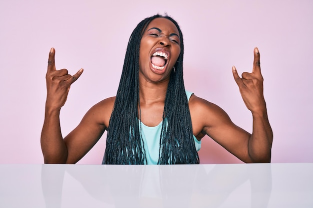 Free photo african american woman with braids wearing casual clothes sitting on the table shouting with crazy expression doing rock symbol with hands up music star heavy concept