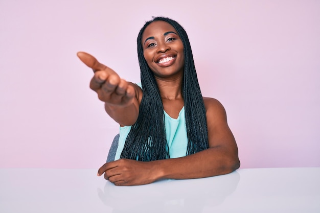 Free photo african american woman with braids wearing casual clothes sitting on the table smiling friendly offering handshake as greeting and welcoming. successful business.