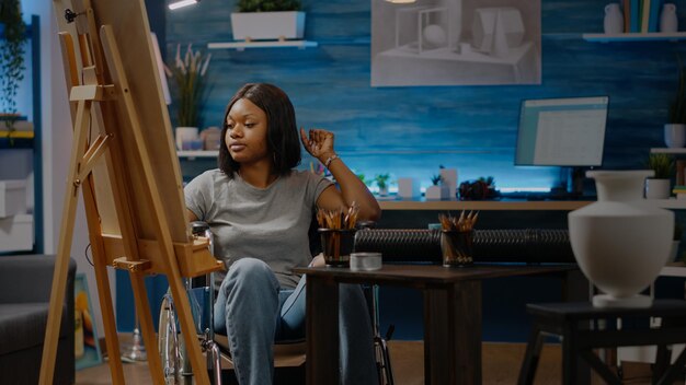 Афро-американская женщина с инвалидом работает над арт-проектом, сидя в пространстве студии. Чернокожий художник-инвалид в инвалидной коляске рисует белый дизайн вазы на холсте и мольберте