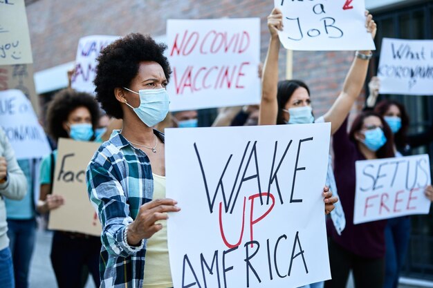 African American woman with protective face mask protesting with crowd of people on city streets and carrying banner with 'wake up America' inscription