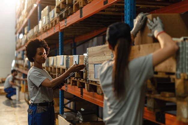 Афроамериканская работница общается со своей коллегой во время подготовки отгрузки на промышленном складе