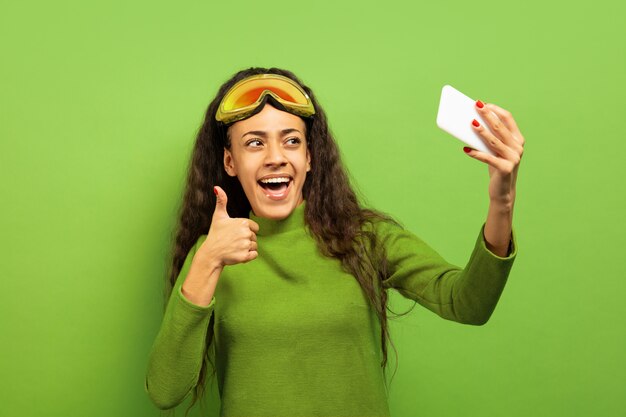 African-american young brunette woman's portrait in ski mask on green studio background. Concept of human emotions, facial expression, sales, ad, winter sport and holidays. Making selfie or vlog.