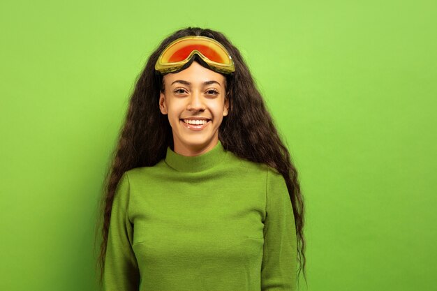 緑のスタジオの背景にスキーマスクでアフリカ系アメリカ人の若いブルネットの女性の肖像画。人間の感情、顔の表情、販売、広告、ウィンタースポーツ、休日の概念。笑顔で、幸せそうに見えます。