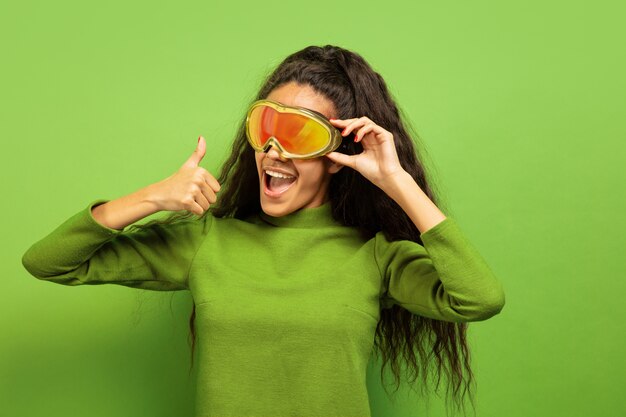 African-american young brunette woman's portrait in ski mask on green studio background. Concept of human emotions, facial expression, sales, ad, winter sport and holidays. Smiling, thumb up.