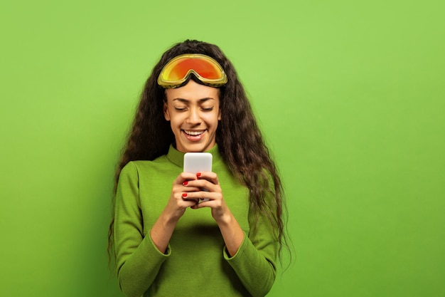 Foto gratuita ritratto di giovane donna bruna afro-americana in passamontagna su sfondo verde studio. concetto di emozioni umane, espressione facciale, vendite, pubblicità, sport invernali e vacanze. utilizzando smartphone.