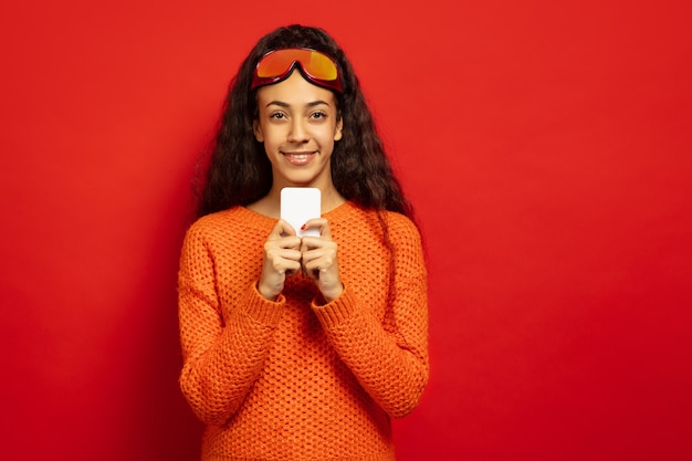 African-american young brunette woman's portrait in ski mask on red studio background. Concept of human emotions, facial expression, sales, ad, winter sport and holidays. Chatting with phone.