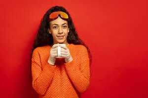 African-american young brunette woman's portrait in ski mask on red studio background. concept of human emotions, facial expression, sales, ad, winter sport and holidays. drinks tea, coffee.