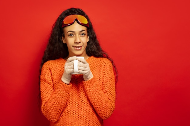 African-american young brunette woman's portrait in ski mask on red studio background. Concept of human emotions, facial expression, sales, ad, winter sport and holidays. Drinks tea, coffee.